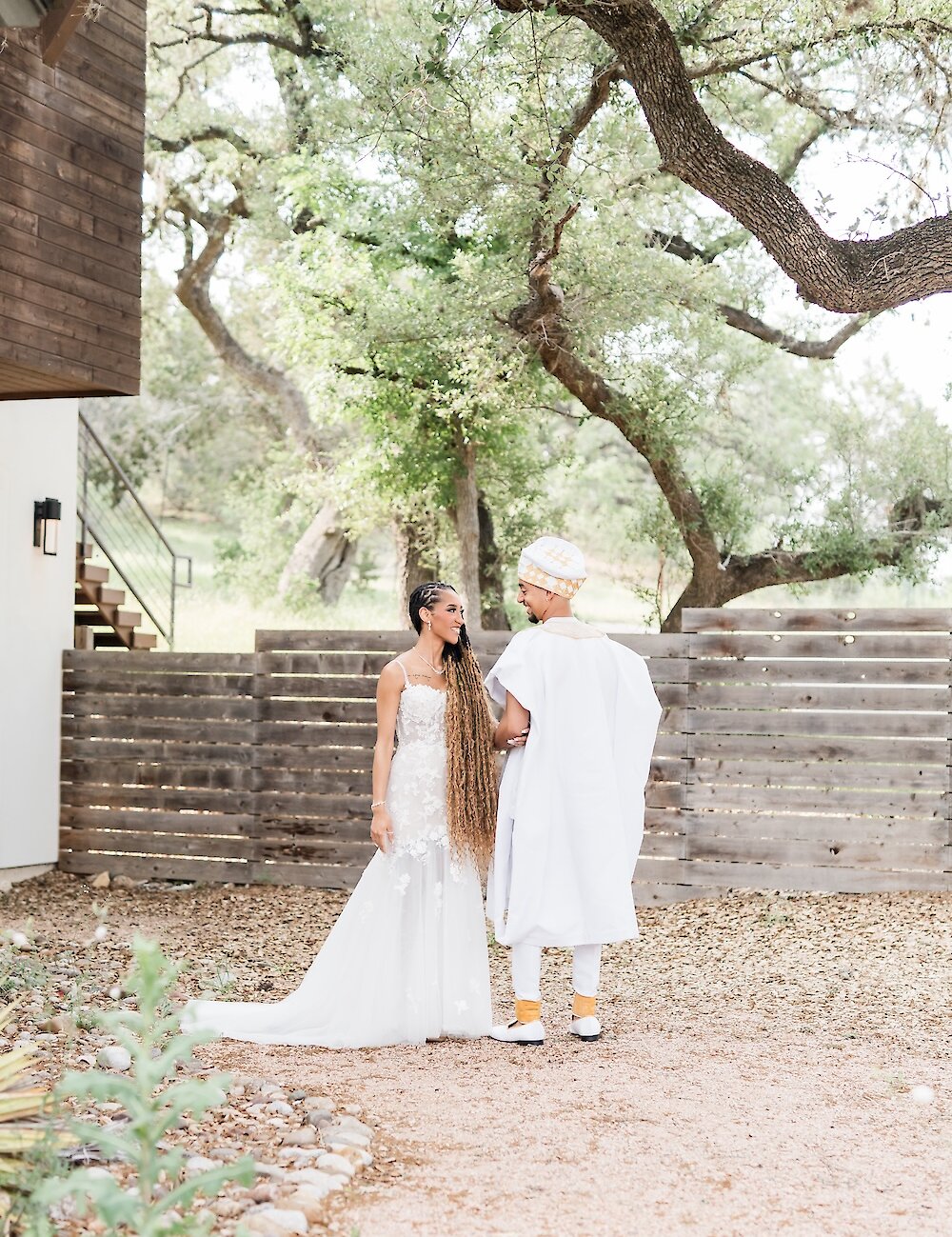 The bride and groom pose facing each other outdoors, with the groom in white and gold traditional attire and the bride in her white lace gown, near a wooden fence.