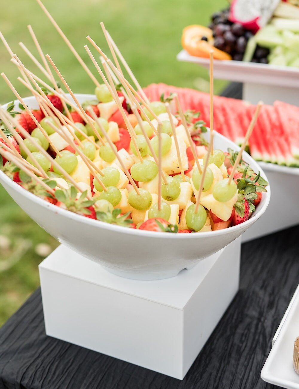 A large white bowl holds fruit salad with toothpicks inserted into pieces of pineapple and green grapes, set on a black linen table for the buffet.
