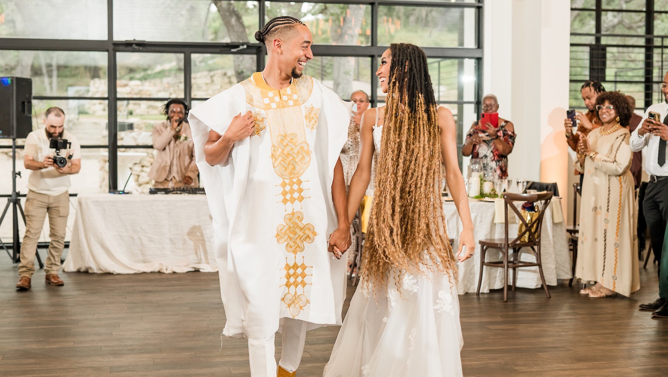 The bride and groom smile, holding hands and wearing white and gold attire, making their grand entrance into the reception hall with cheering guests.