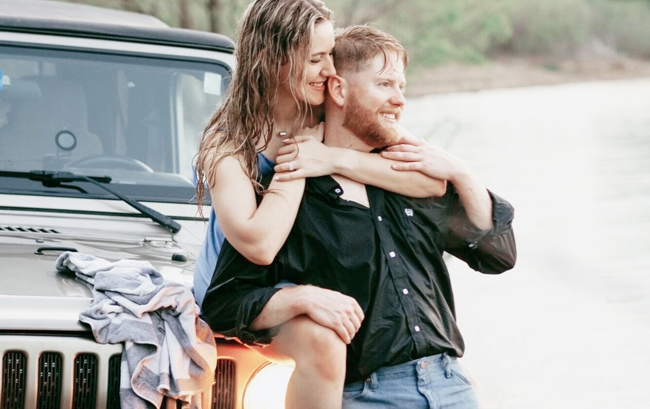 A happy engaged couple embraces near the water, with the woman sitting on the hood of a Jeep while hugging the man from behind for a casual outdoor portrait.