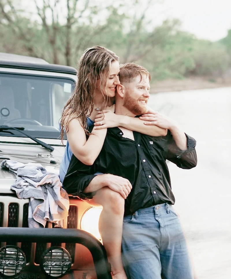 A happy engaged couple embraces near the water, with the woman sitting on the hood of a Jeep while hugging the man from behind for a casual outdoor portrait.