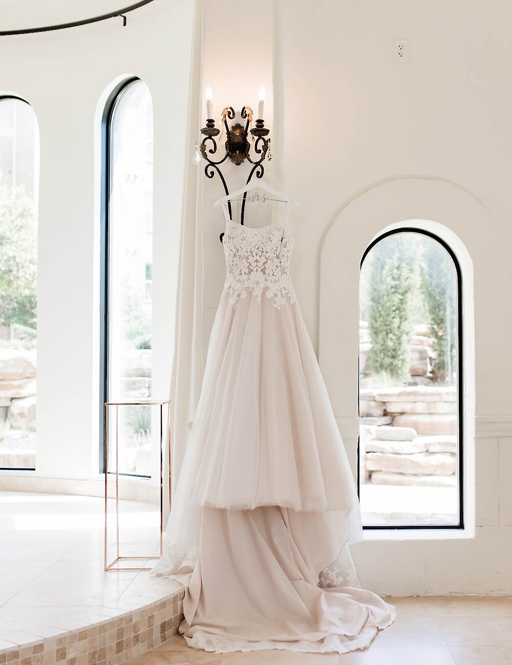 A beautiful blush and white wedding dress with a lace bodice hangs from a sconce on a white wall, framed by tall arched windows in a bright chapel.