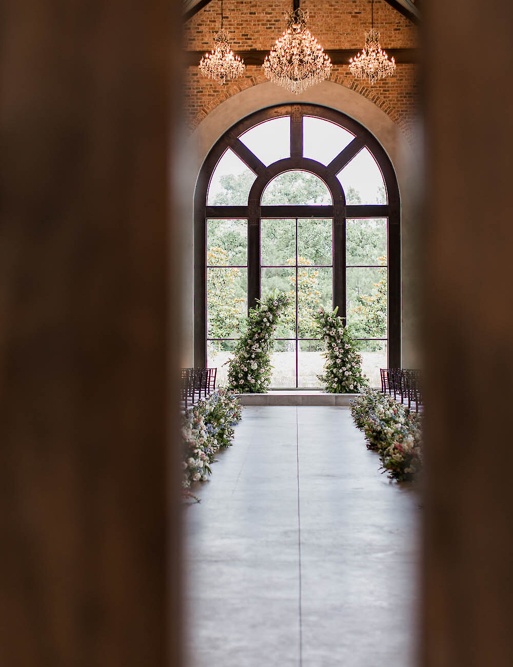 An empty industrial-style ceremony space featuring a towering arched window, brick walls, crystal chandeliers, and an aisle lined with low floral arrangements.
