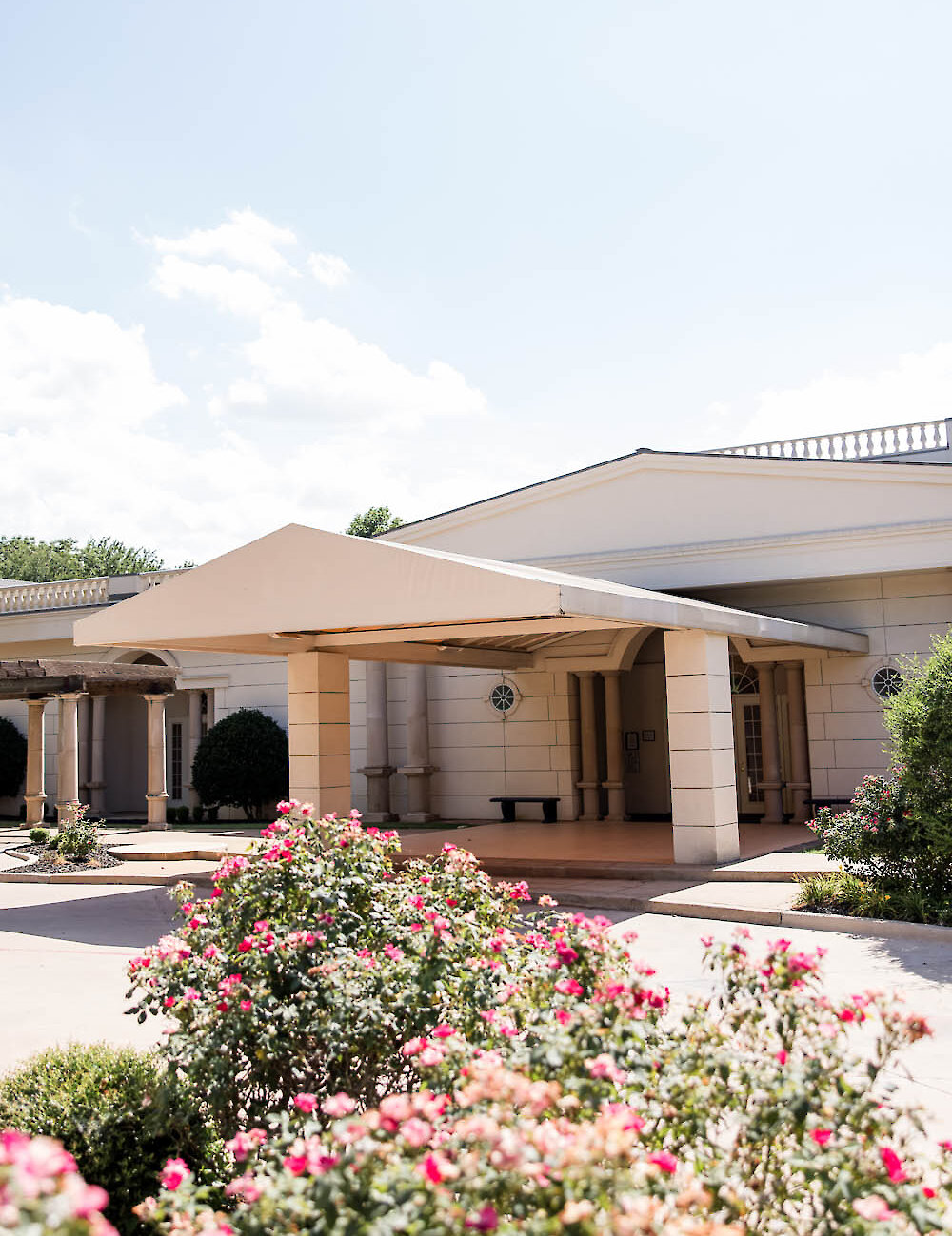 The exterior of a Southern-style wedding venue featuring a large covered entrance porte-cochère, white columns, and bright pink rose bushes in the foreground.