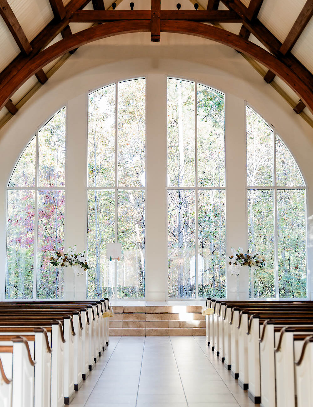A bright chapel interior featuring vaulted wooden trusses, white pews lining the aisle, and tall arched windows overlooking autumn trees.