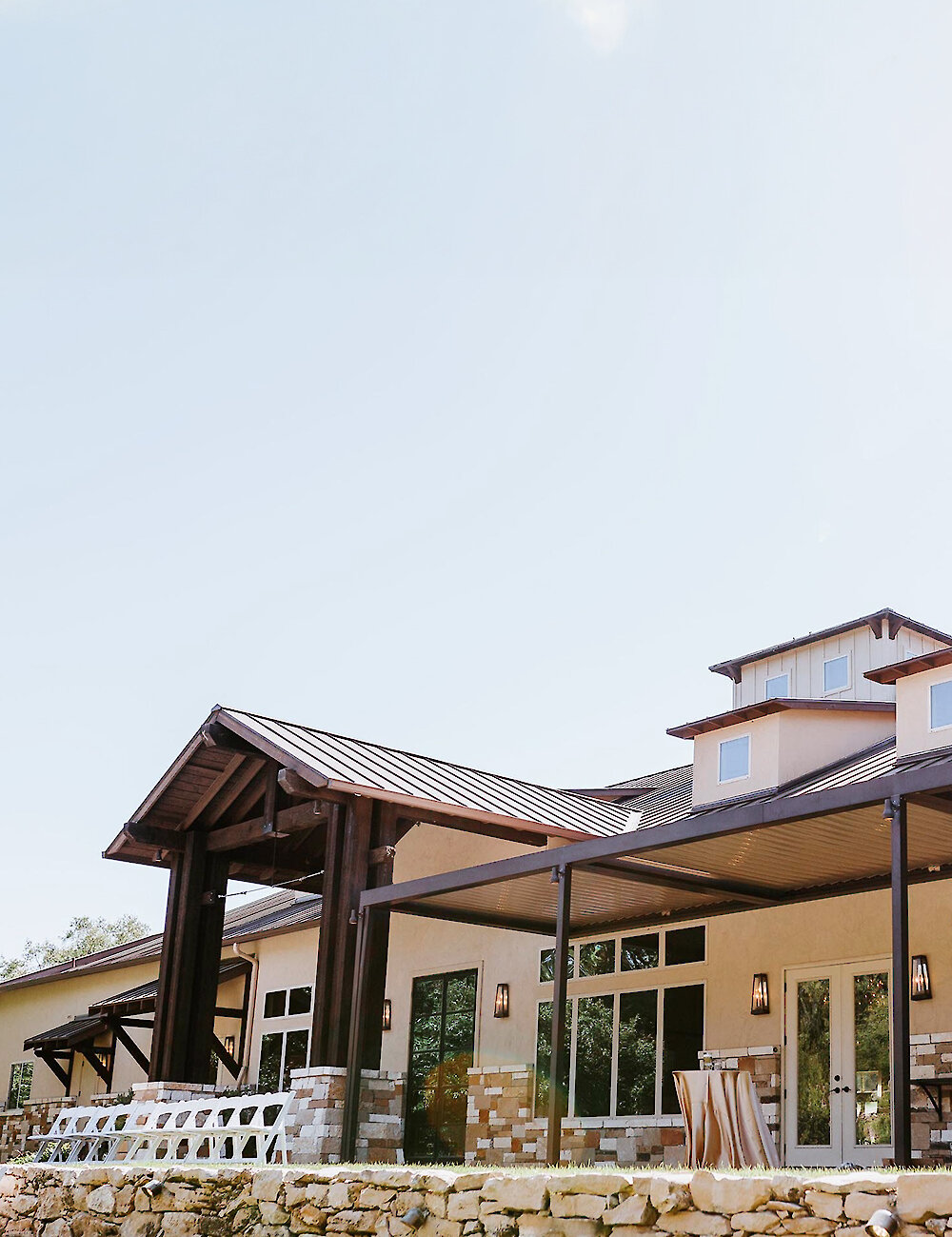 The exterior of a rustic modern venue featuring a beige facade, dark wooden accents, a covered patio, and a retaining wall made of light-colored stacked stone.