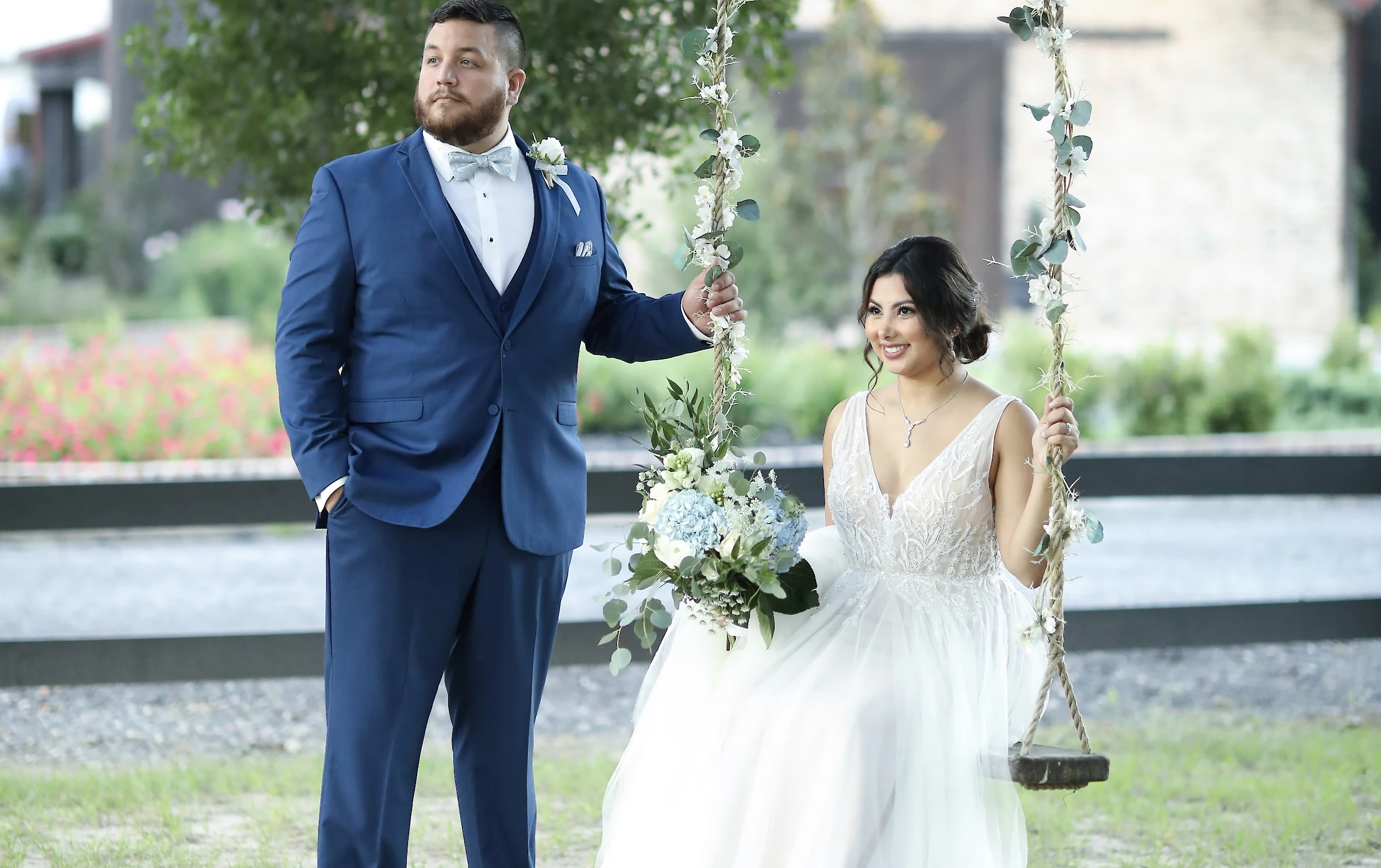 The bride sits on a wooden swing decorated with greenery, holding a blue and white bouquet, next to the groom in a bright blue suit for an outdoor portrait.