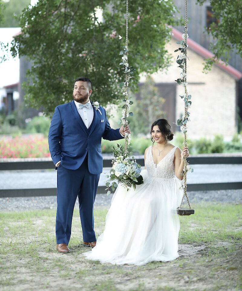 The bride sits on a wooden swing decorated with greenery, holding a blue and white bouquet, next to the groom in a bright blue suit for an outdoor portrait.