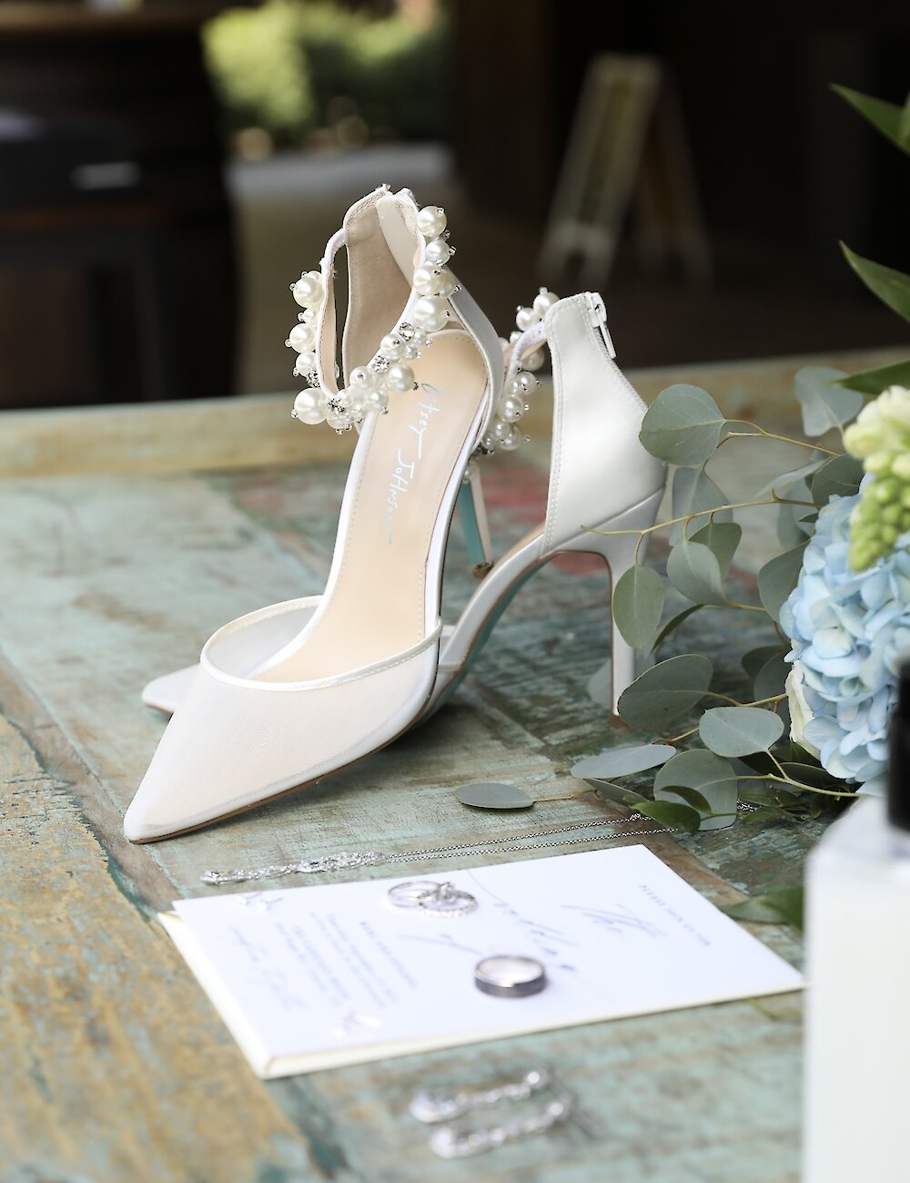 A white pointed bridal pump with a pearl ankle strap sits on a rustic wooden table with the wedding rings, necklace, and blue and green florals.