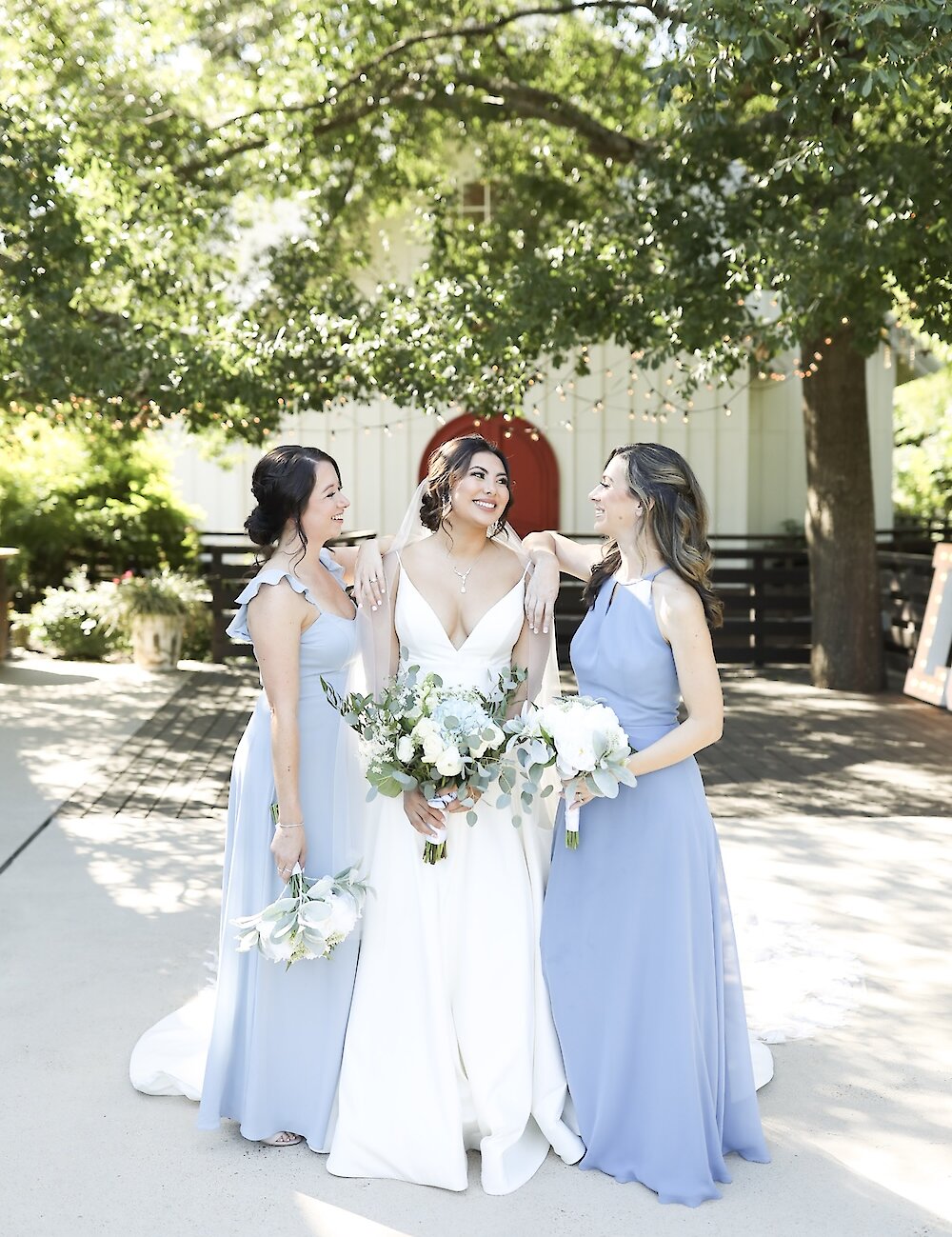 The smiling bride in a white gown poses outdoors with two bridesmaids in floor-length dusty blue dresses, holding bouquets under a large tree.