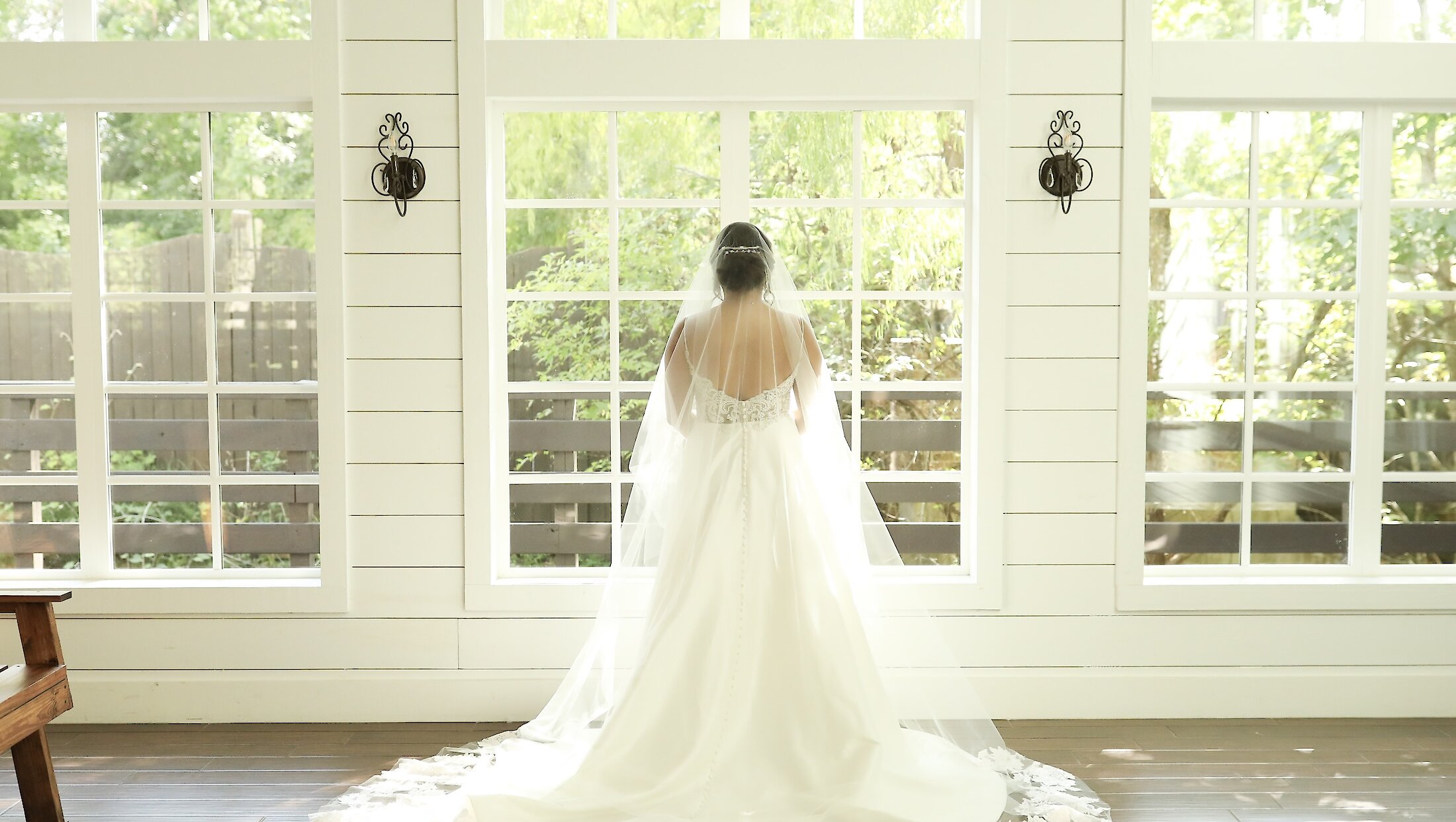 The bride's back is visible, showcasing her open-back gown and long, lace-trimmed veil trailing on the dark wood floor in front of three large sunlit windows.