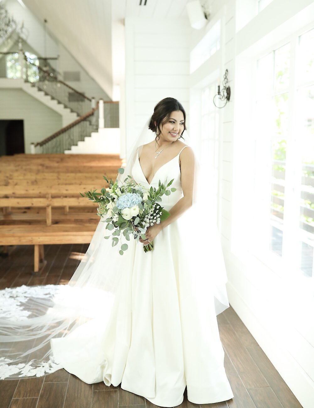 A smiling bride in a white satin gown with a plunging neckline holds a bouquet of white and blue flowers while posing near a bright window inside a white shiplap chapel.