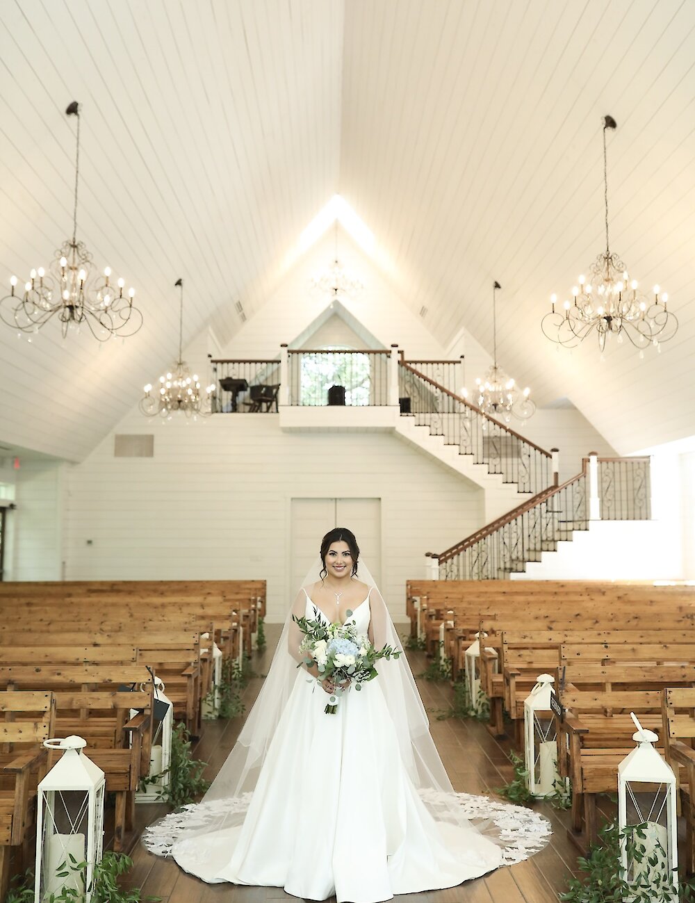 The smiling bride stands centered in the aisle of a white vaulted chapel, wearing a white satin gown and veil, framed by wooden pews and chandeliers.