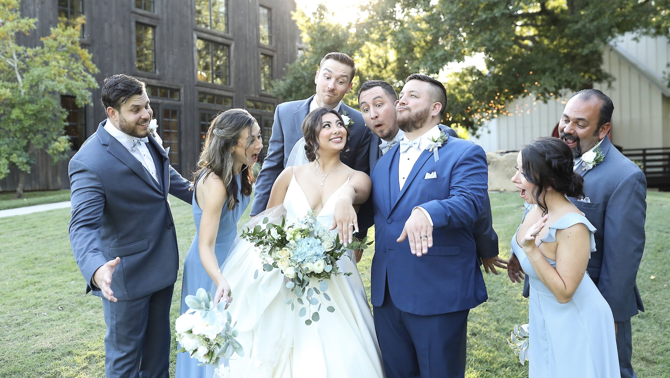 The bride and groom pose for a funny outdoor photo with their wedding party, showing exaggerated shock and surprise in front of a dark wood barn.