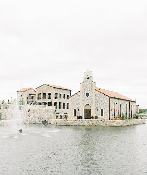Stone chapel with arched windows, wooden double doors, and a small bell tower, surrounded by a stone-lined water feature under a clear blue sky.