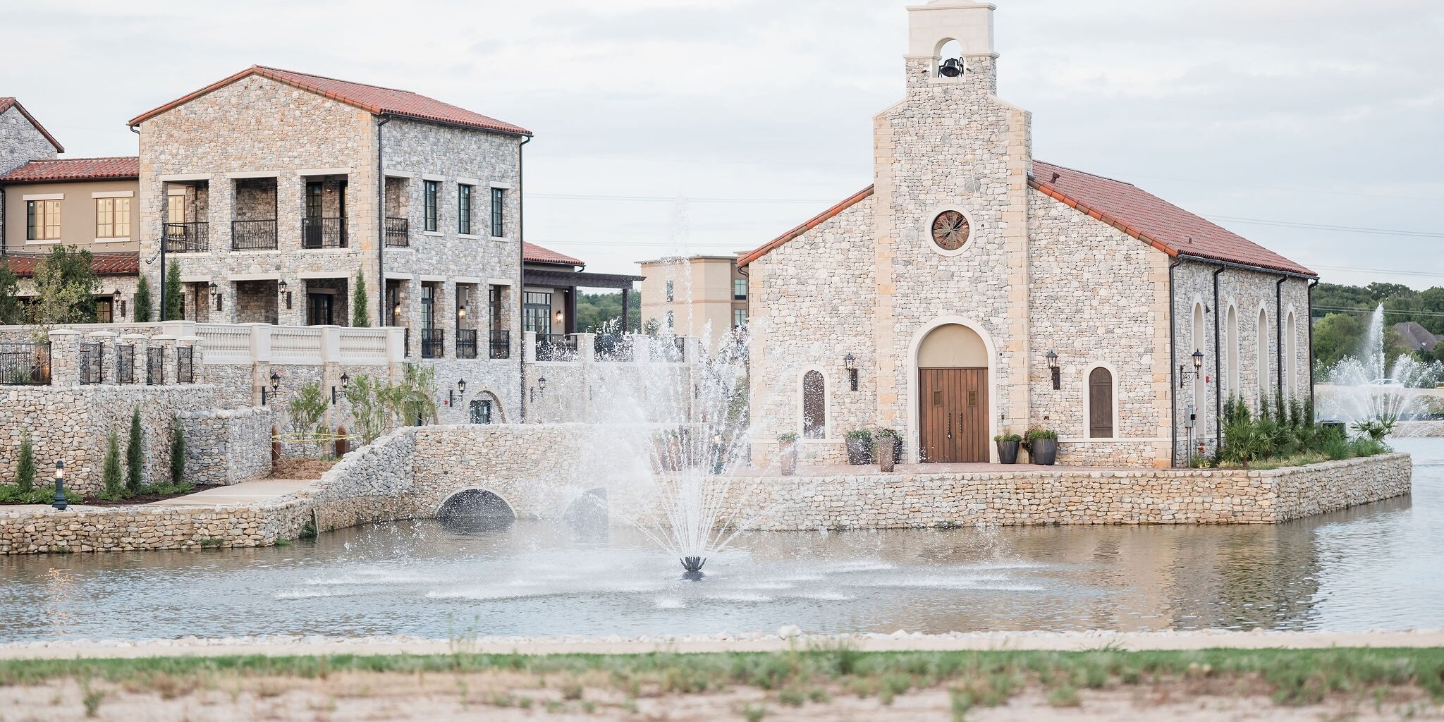 Wide exterior view of a Mediterranean-style wedding venue complex featuring a stone chapel with a bell tower and a multi-story reception building, all situated on a pond with a central water fountain.