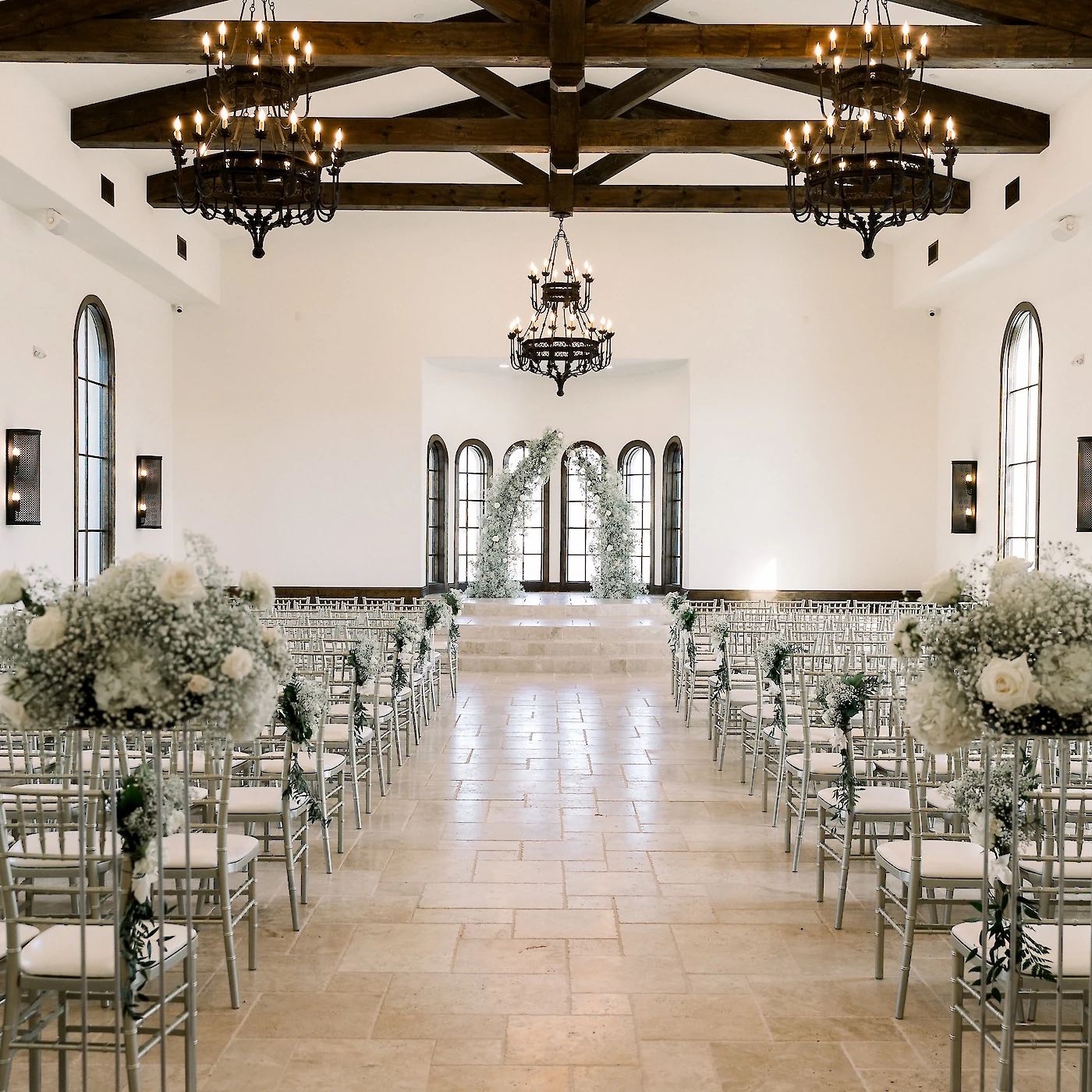 Elegant wedding ceremony hall featuring a high white vaulted ceiling with dramatic exposed wooden trusses and dark iron chandeliers. The aisle is lined with silver chiavari chairs and tall floral arrangements of white roses and baby's breath, leading to a trio of arched windows and a floral arch altar.