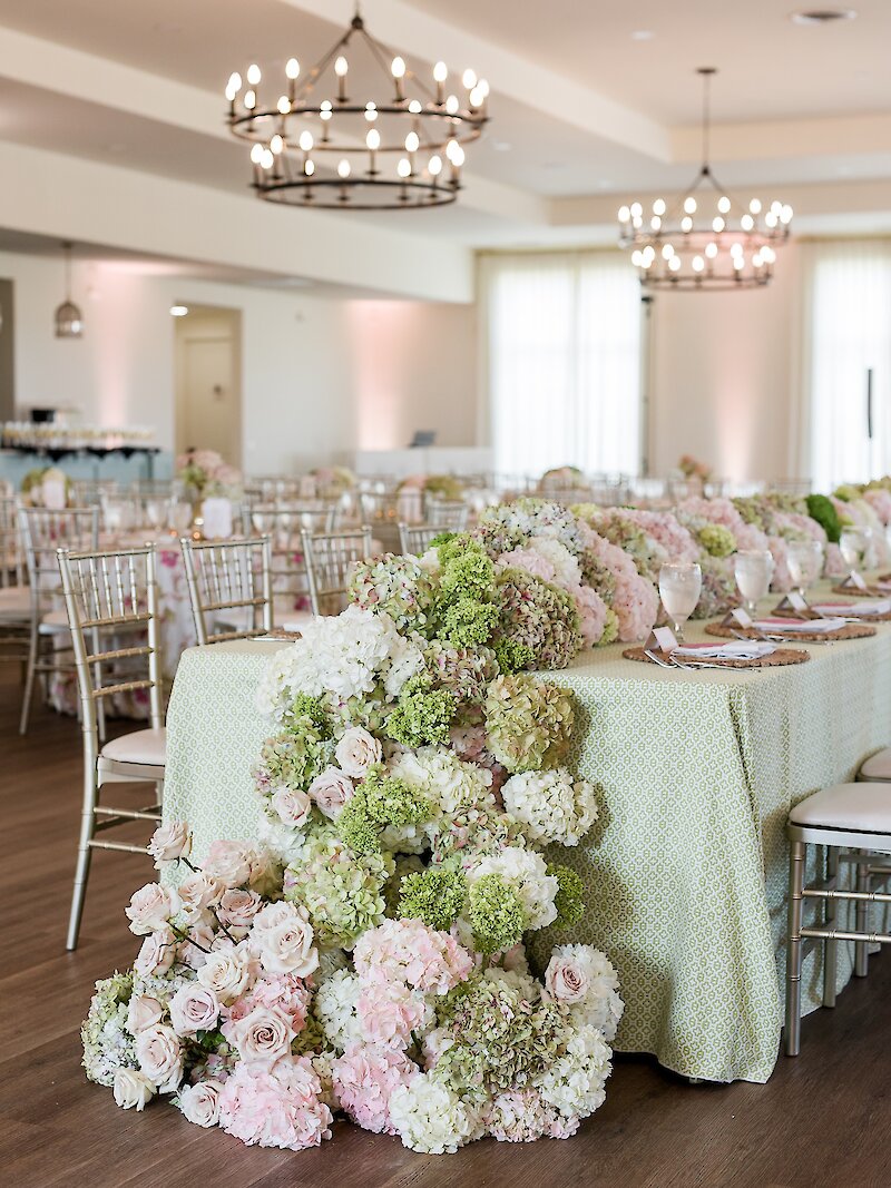 Luxurious wedding reception head table covered in a sage green patterned tablecloth, draped with an abundant garland of pink and green hydrangeas and blush roses, set in a ballroom with wagon wheel chandeliers and gold chiavari chairs.