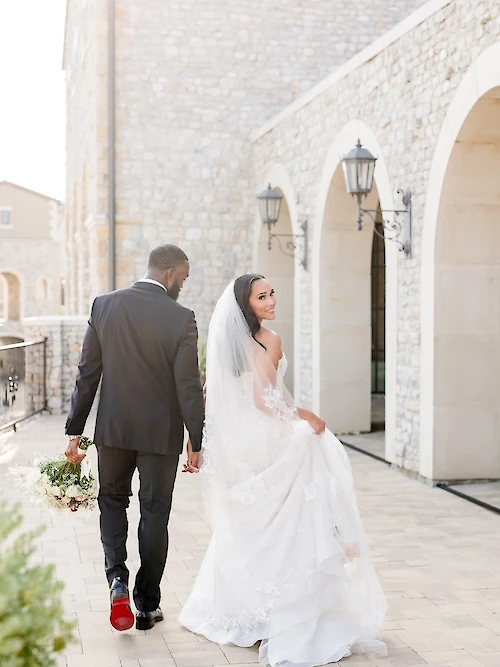 Sunlit wedding portrait of a Black couple walking hand-in-hand down a stone courtyard lined with arched walkways. The bride smiles back, showing off her lace gown and veil, while the groom's red shoe sole is visible.