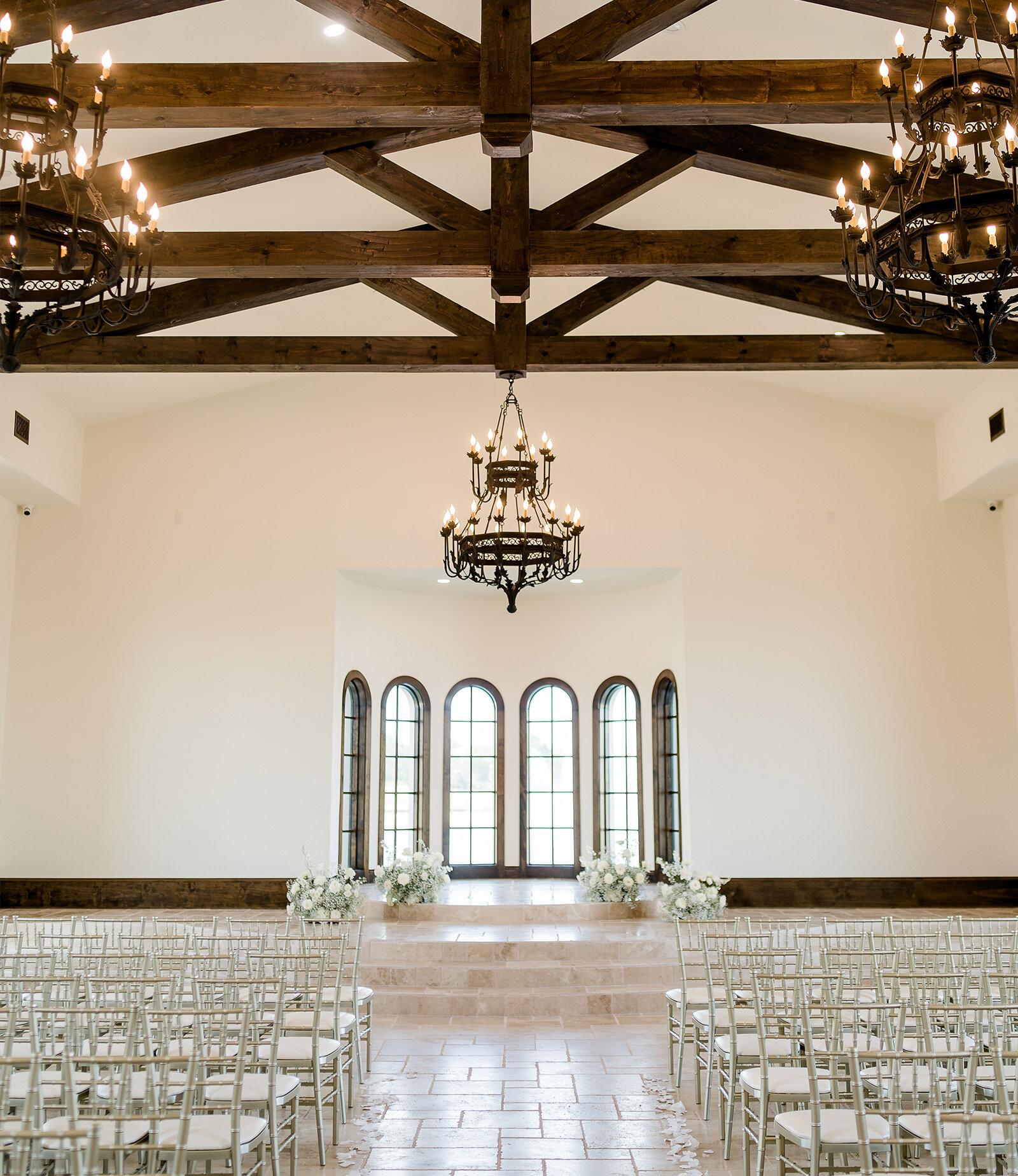 Modern wedding reception ballroom featuring tables set with white linens, silver Chiavari chairs, and light green accent plates. The room is illuminated by large iron chandeliers heavily draped with lush green vines and foliage.