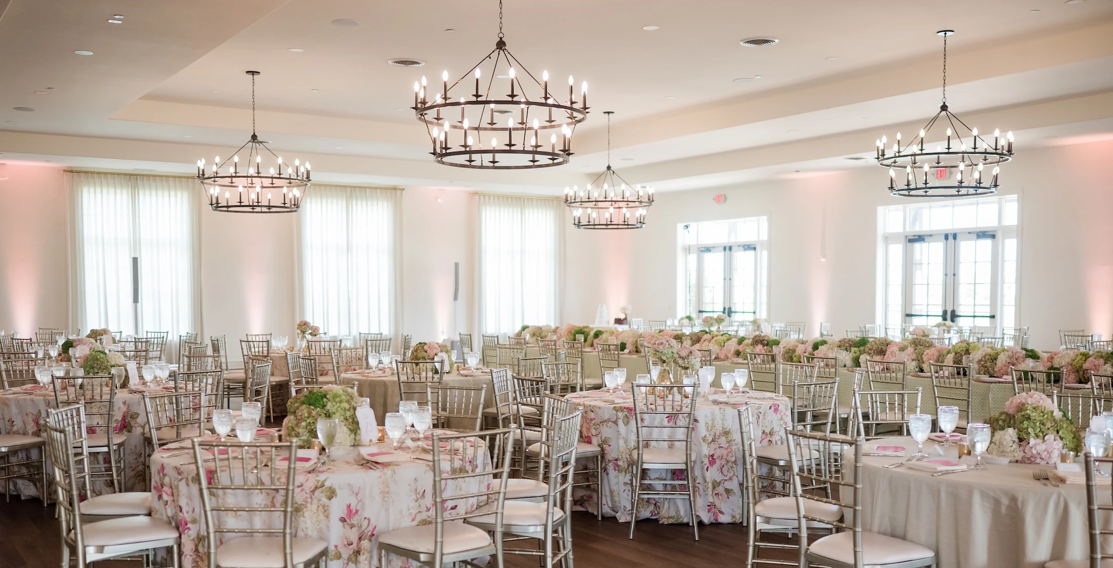 A bright, elegant ballroom set for a reception with round tables featuring floral and beige linens, chiavari chairs, and large chandelier lighting.
