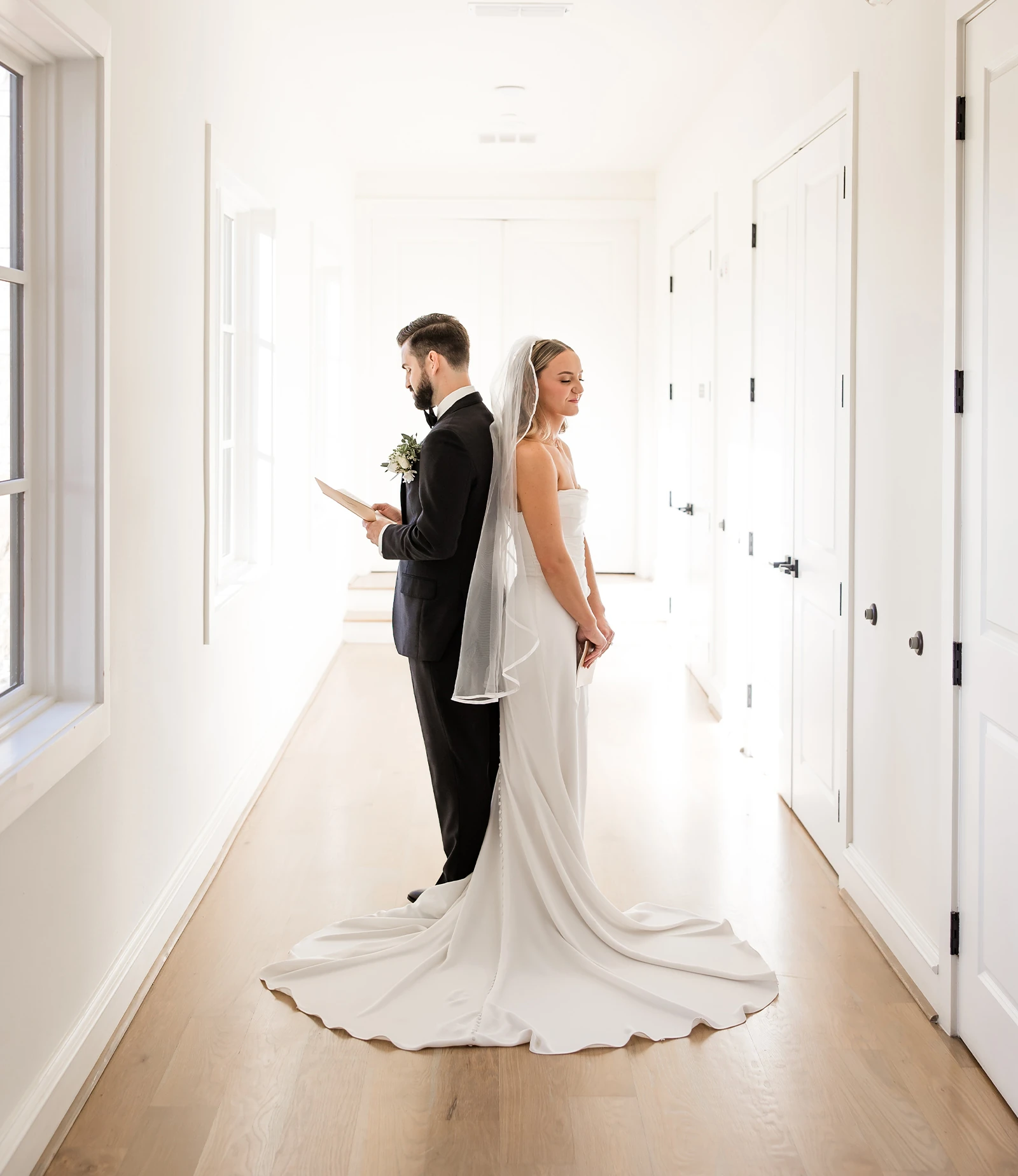 Grand and luminous wedding ceremony hall featuring a vaulted white ceiling with heavy exposed wooden beams and large iron chandeliers. The room is set with rows of white chiavari chairs facing a raised altar with arched windows.