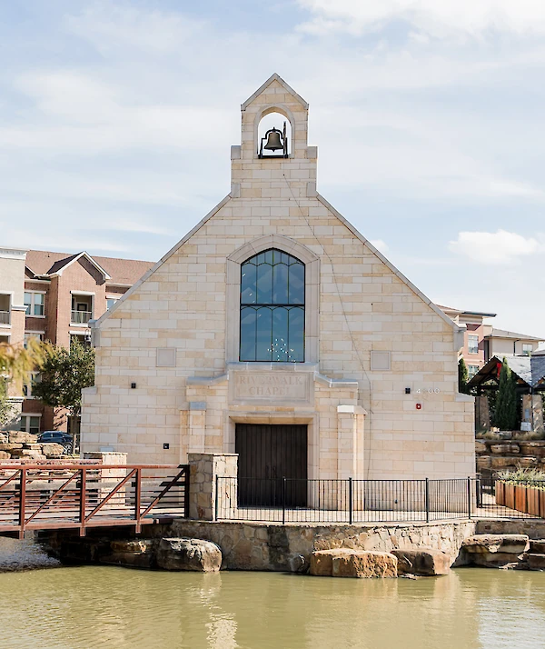 The Riverwalk Chapel, a stone building with a large arched window and a bell in the steeple, viewed across a small pond.