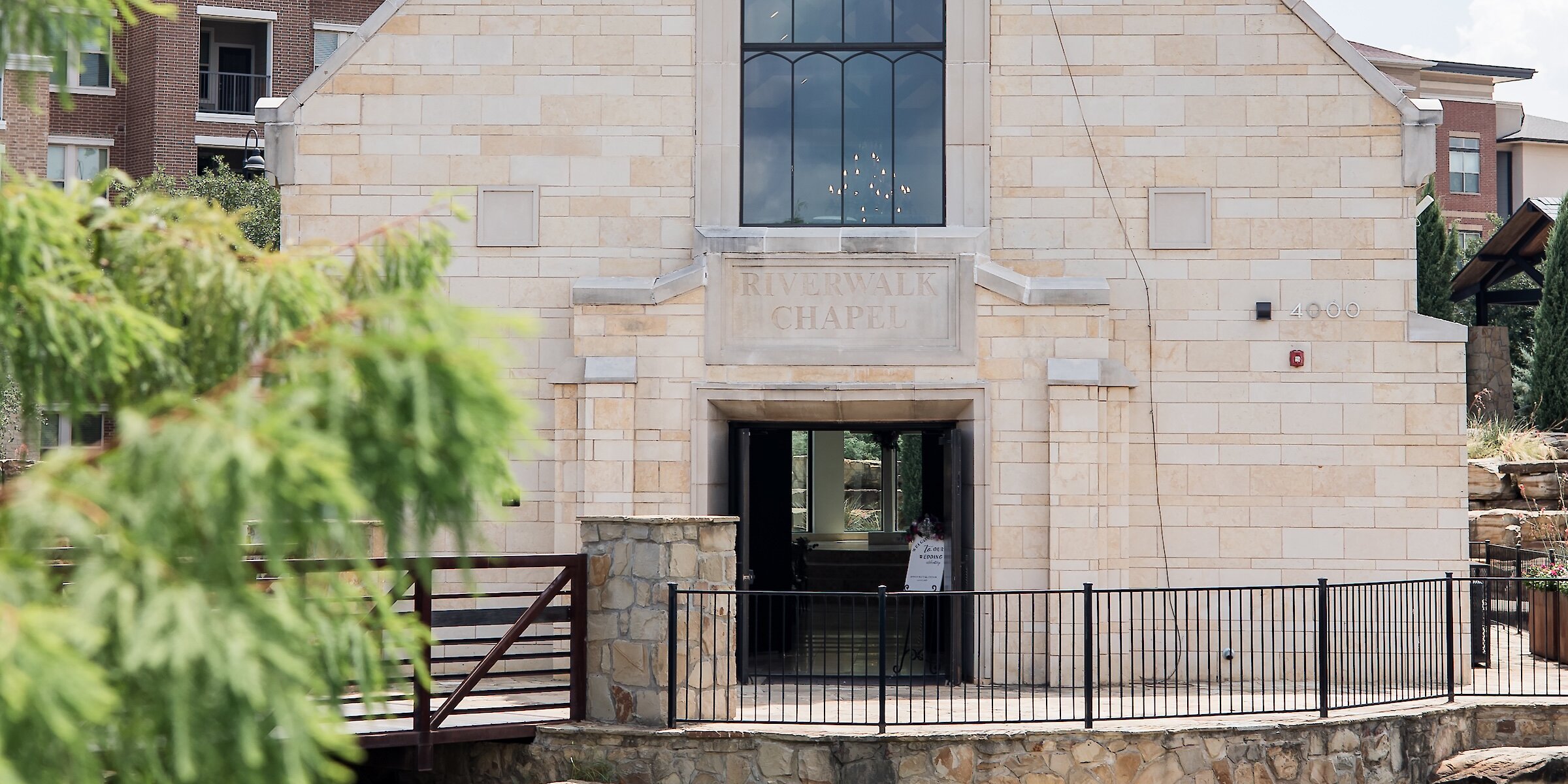 EThe Riverwalk Chapel, a stone building with a large arched window and a bell in the steeple, viewed across a small pond.