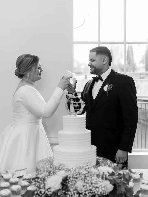 Classic black and white wedding photo of the bride and groom toasting with champagne flutes over their tiered white wedding cake. The groom wears a black tuxedo, and the bride wears a long-sleeved satin gown.