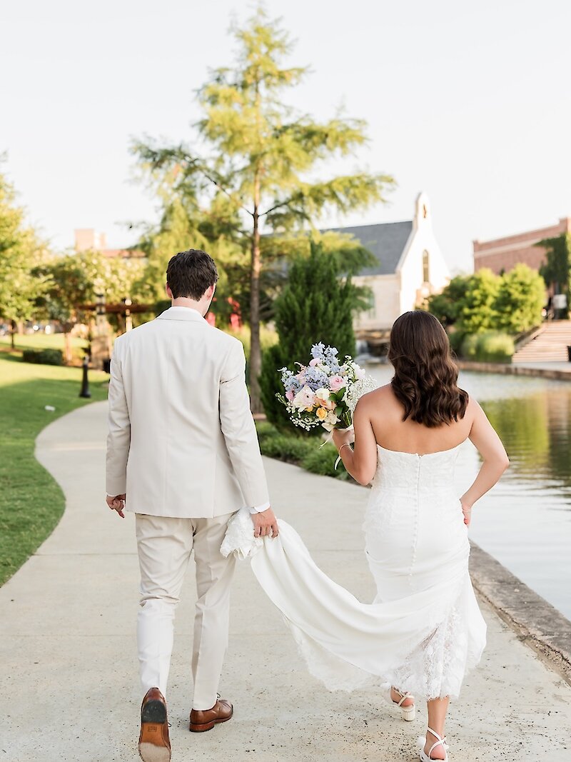Back view of the newlywed couple walking along a path next to a water feature with a small chapel visible in the background. The groom wears a light beige suit, and the bride wears a strapless lace gown and carries a pastel floral bouquet.