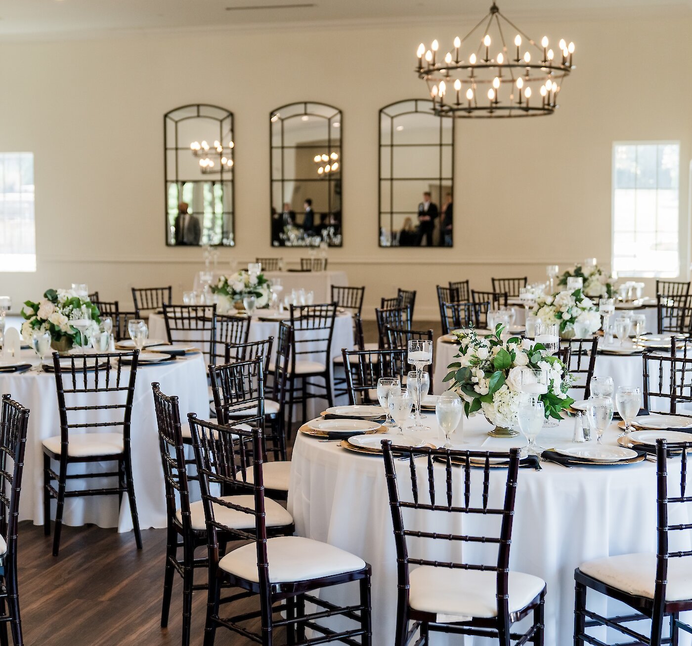 Elegant wedding reception ballroom featuring white tablecloths, dark wood Chiavari chairs, and gold charger plates. Tables are decorated with lush white and green floral centerpieces, with three tall, arched black mirrors reflecting the room.