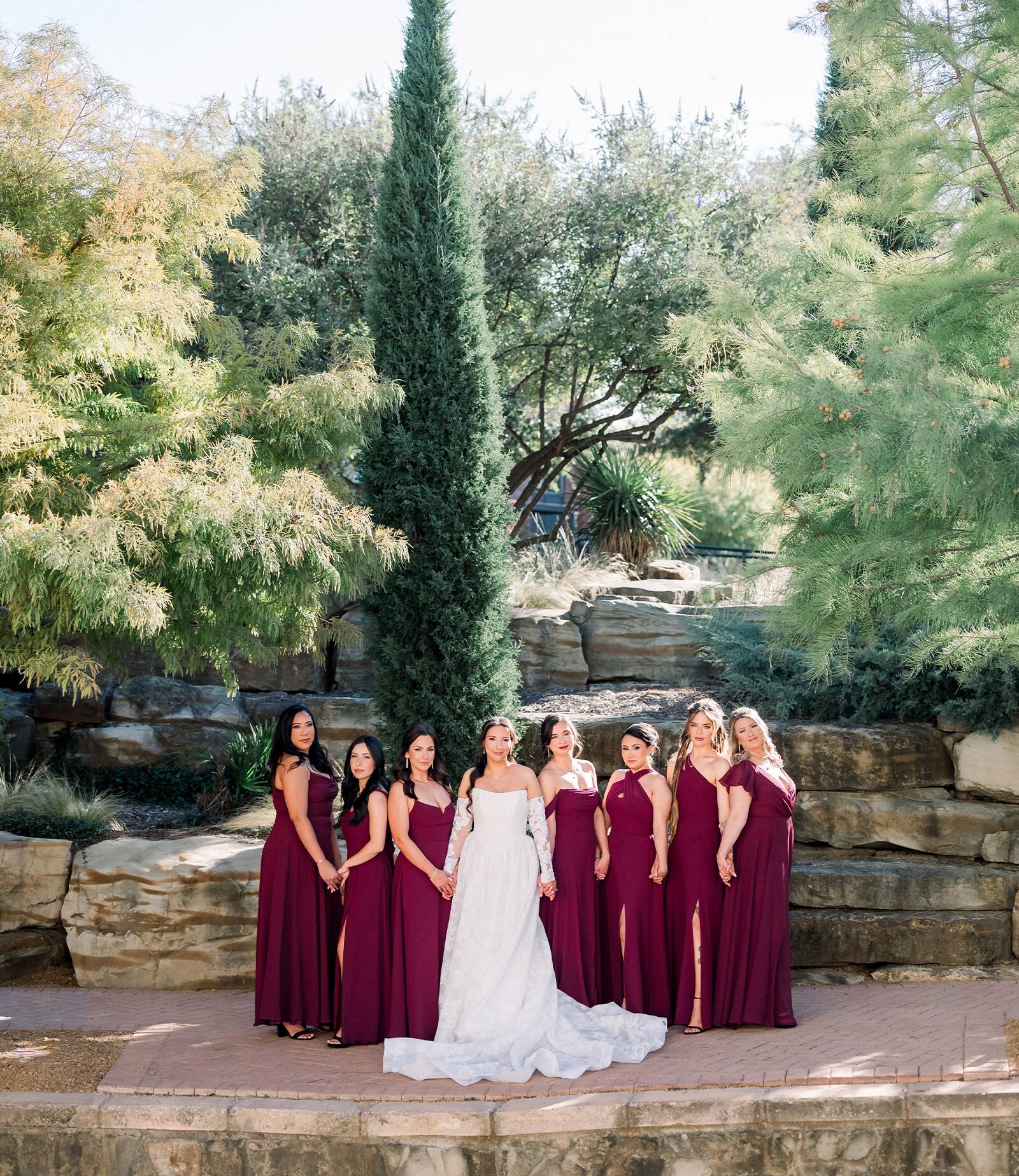The bride and groom pose lovingly on a stone patio in front of a layered, natural rock waterfall and pond, with the bride's veil sweeping the ground.