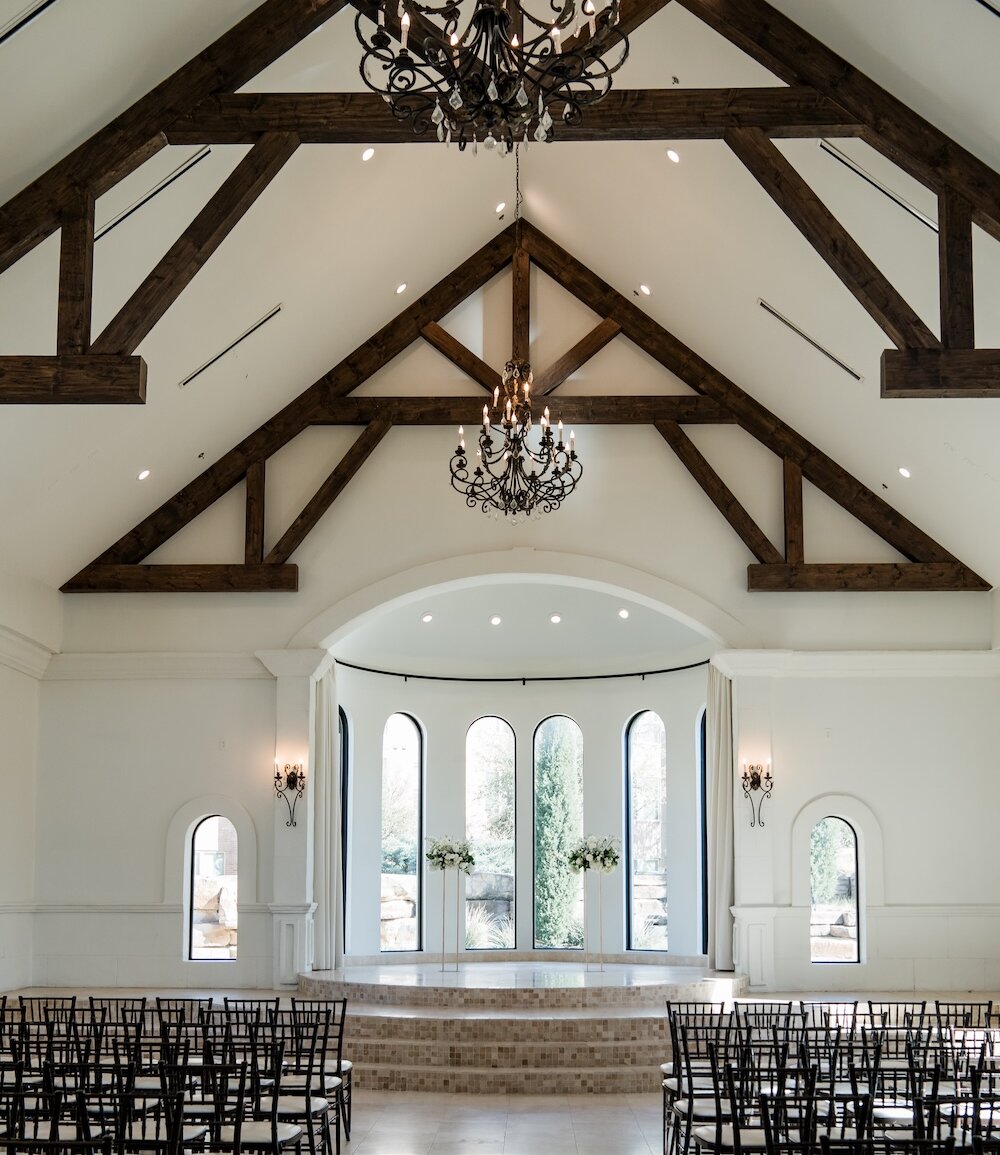 The bright interior of a wedding chapel featuring a vaulted ceiling with dark wood beams, arched windows, and white floral arrangements.