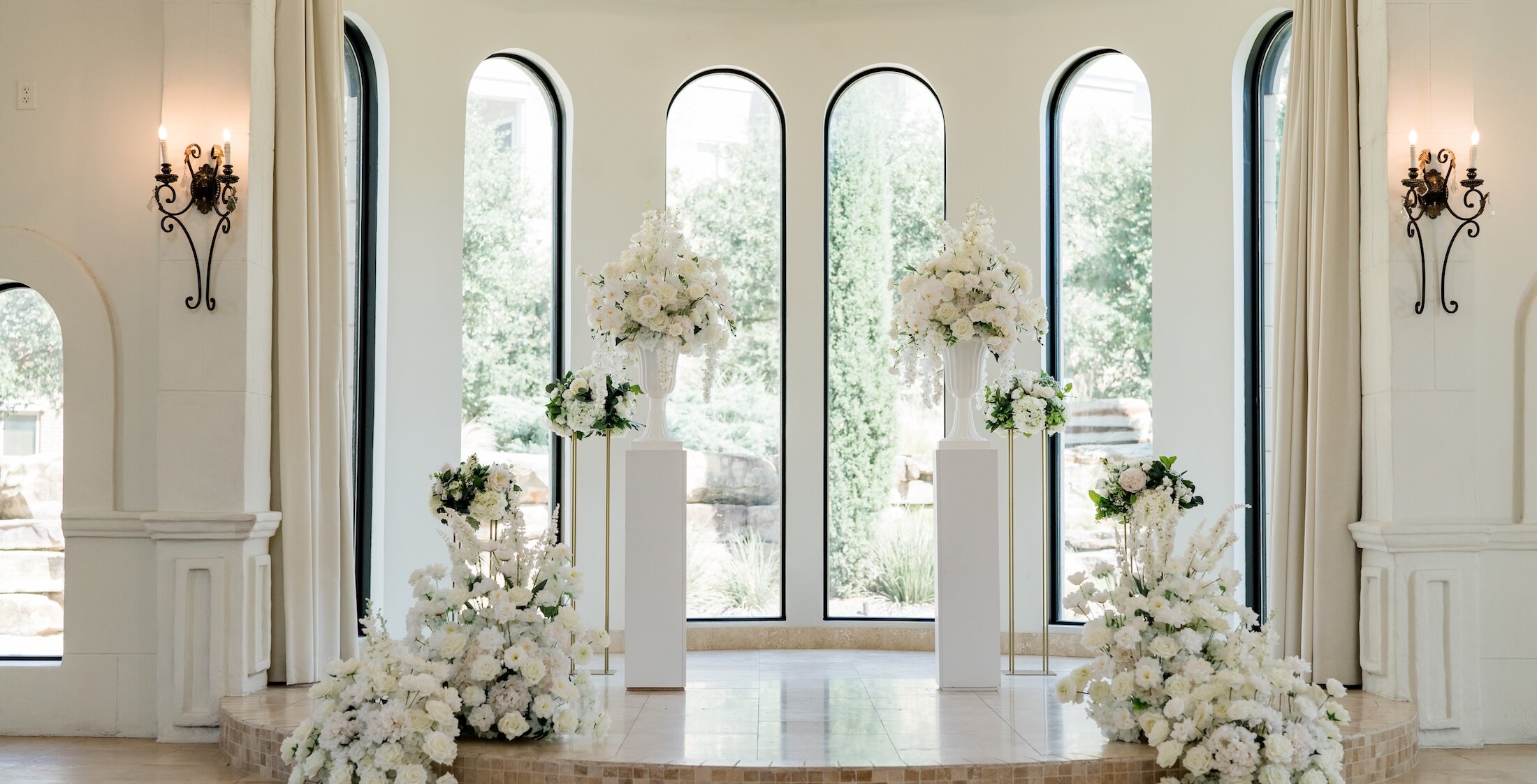 Elegant wedding ceremony altar featuring a curved white platform backed by three tall arched windows. The space is decorated with lush arrangements of white flowers on white and gold pedestals, framed by white curtains and sconces.