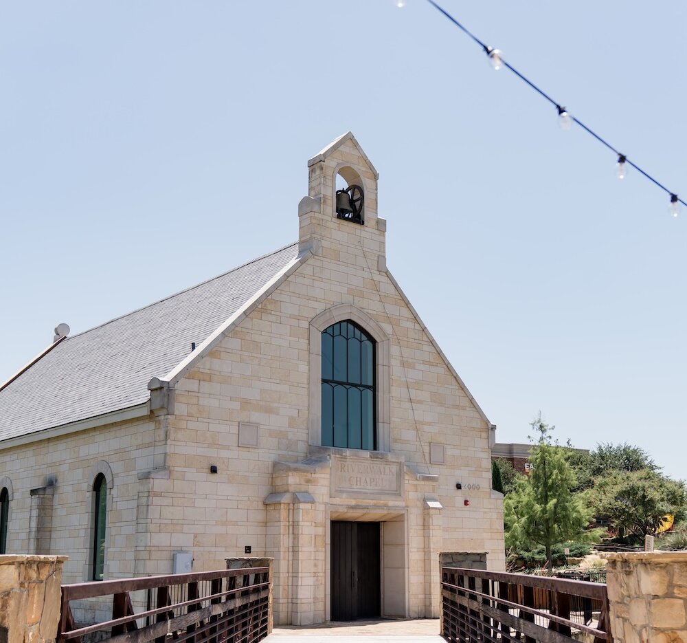 Exterior of the Riverwalk Chapel featuring a light stone facade, bell tower, and large arched window, viewed down a wooden bridge with rustic metal railings, under a bright sky with string lights overhead.