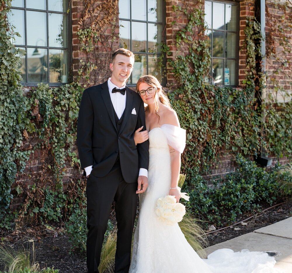 Wedding portrait of the couple posing outside a rustic brick building covered in ivy, with large black-framed windows. The groom wears a classic black tuxedo, and the bride wears an off-the-shoulder lace gown, leaning on the groom's shoulder and holding a white rose bouquet.