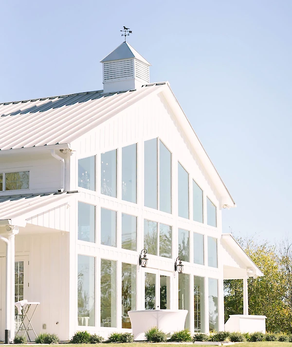 White barn-style building with vertical siding, black shutters, a metal roof, and a cupola, surrounded by neatly trimmed shrubs and green lawn under a partly cloudy sky.