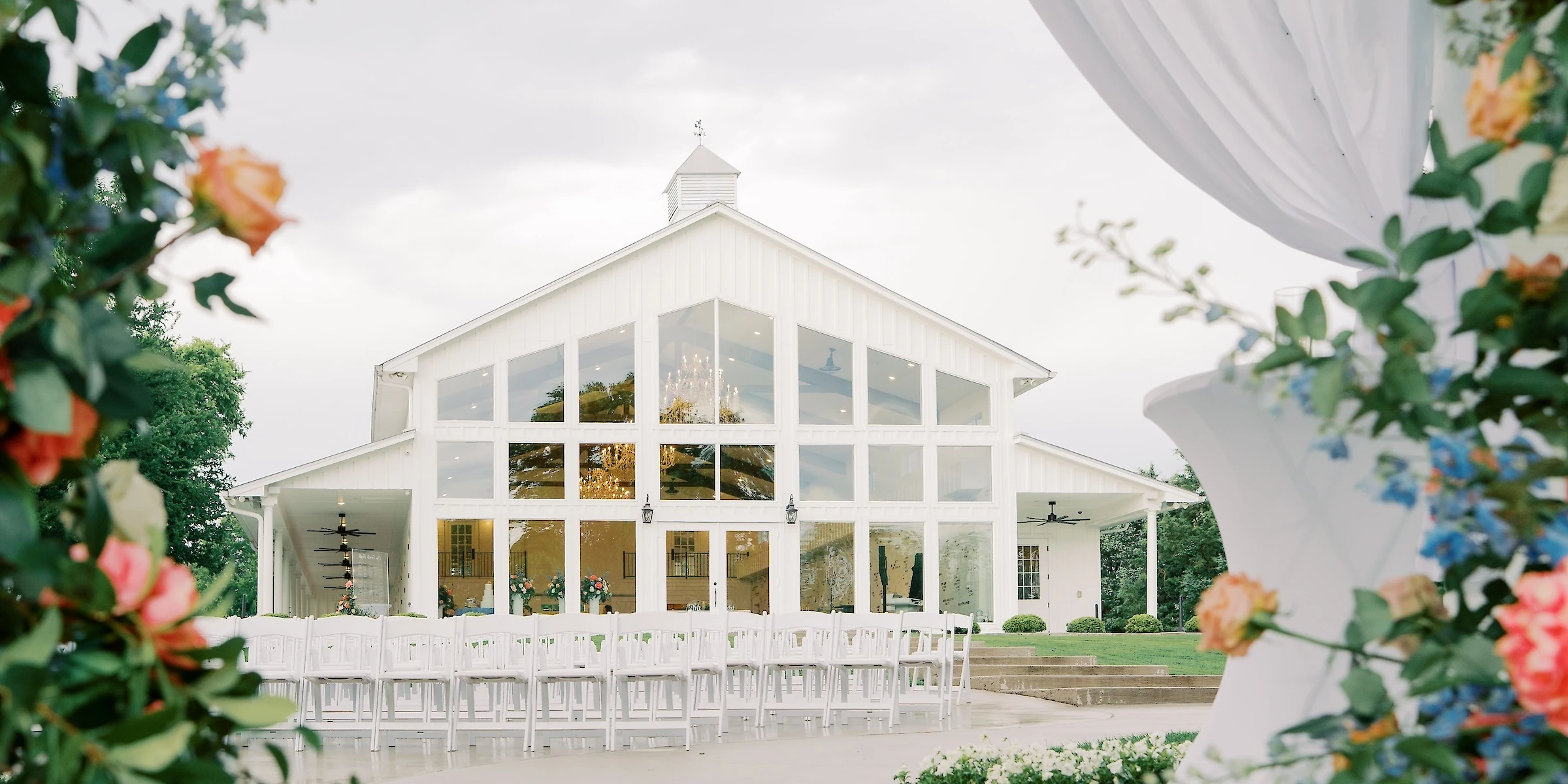 An outdoor wedding ceremony site with white chairs facing a modern white barn venue with large glass windows, framed by sheer fabric and flowers.