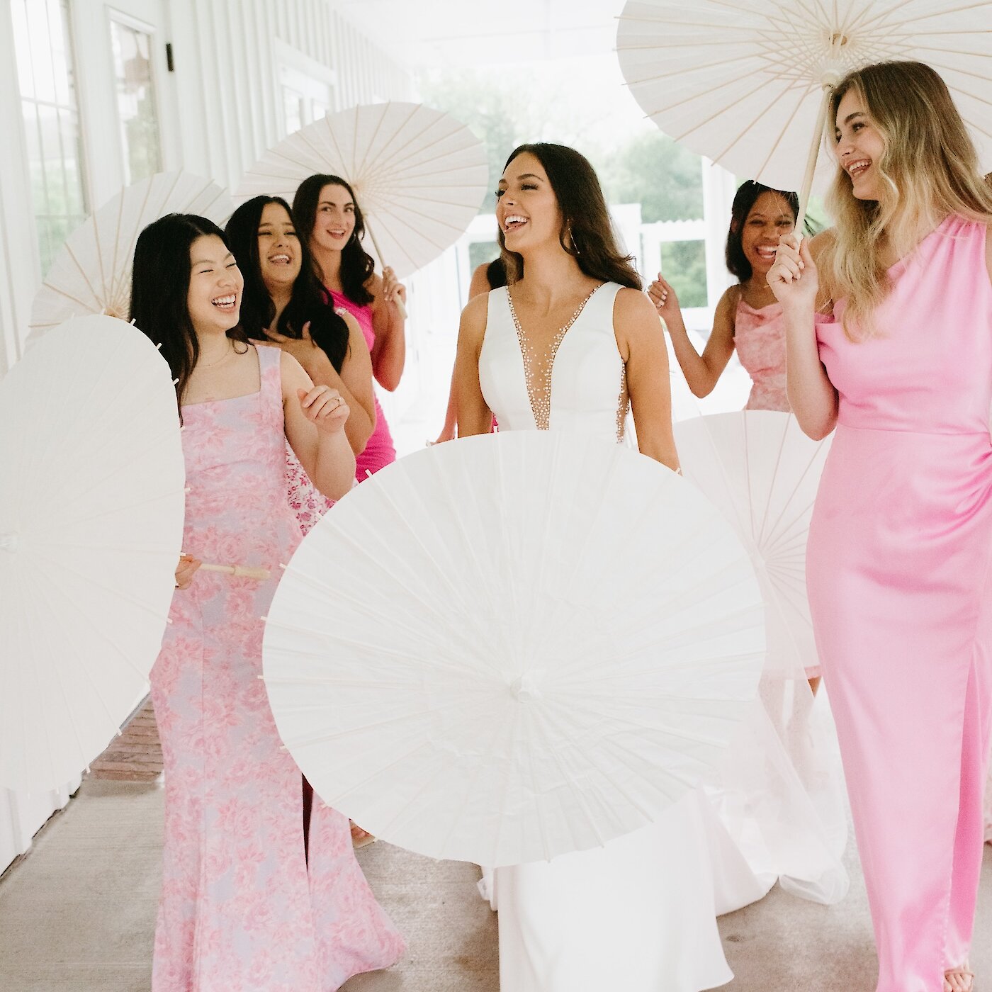Joyful bridal party photo with the bride and bridesmaids walking and laughing on a covered porch. The bride wears a sleek white gown, and the bridesmaids wear varied pink dresses, all carrying white paper parasols.