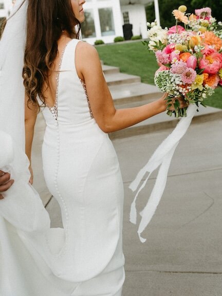 Close-up back view of a bride in a fitted sleeveless white gown with a row of buttons and beaded trim along the open back, holding a large, vibrant bouquet of pink, coral, and yellow flowers with long trailing ribbons.
