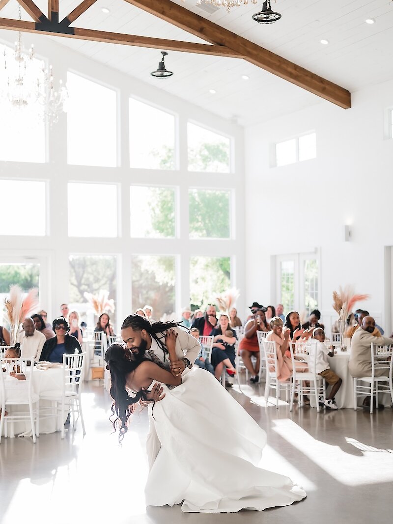 Joyful, sunlit wedding first dance moment with the groom dipping the bride in a bright, modern farmhouse reception hall. The room features a high vaulted ceiling with exposed wooden trusses and a wall of large windows.