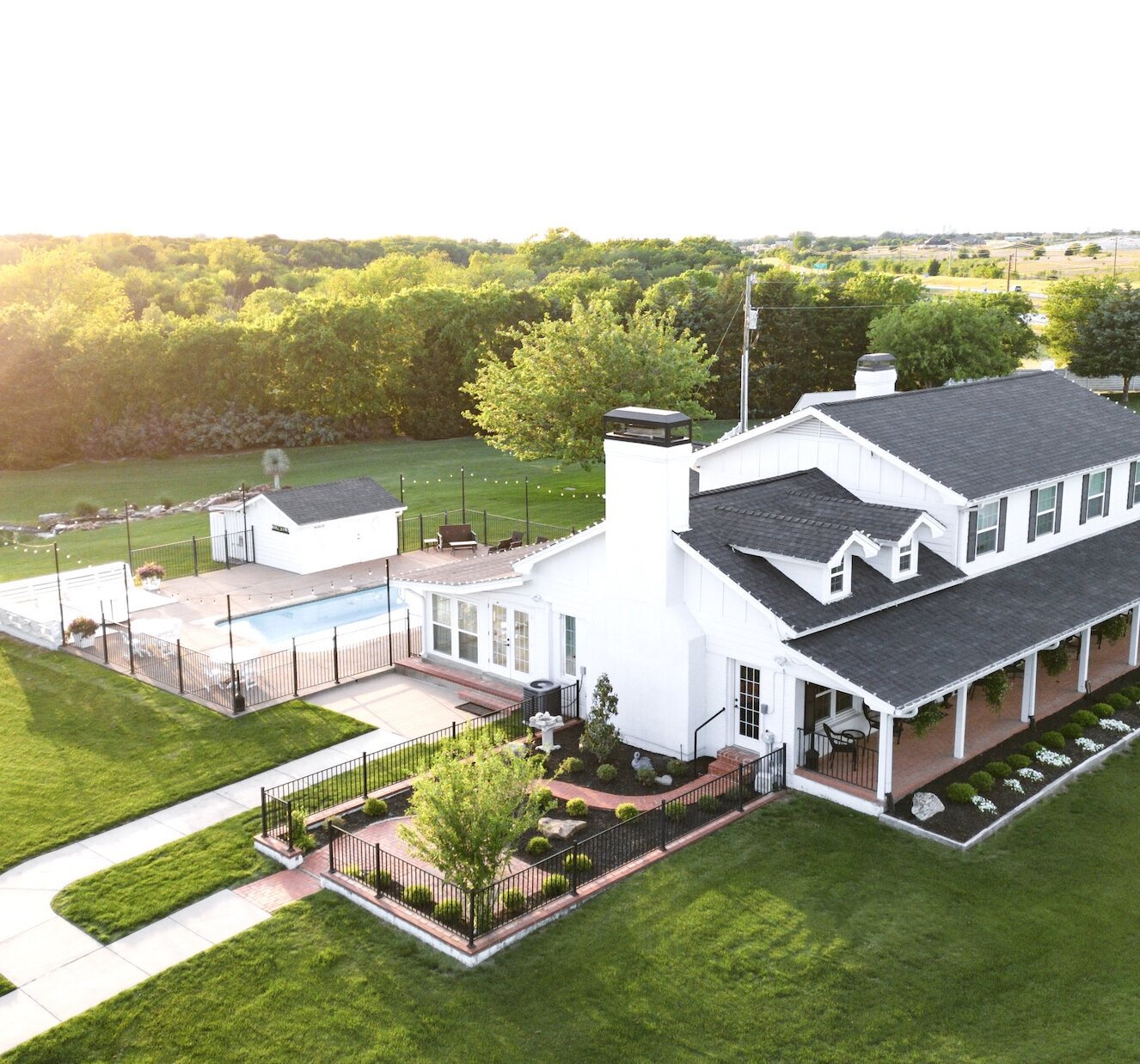 Aerial view of a white farmhouse wedding venue with a black roof and large covered porch, situated on a green lawn. The property includes a swimming pool, winding walkway, and wooded area with a pond visible during sunset.