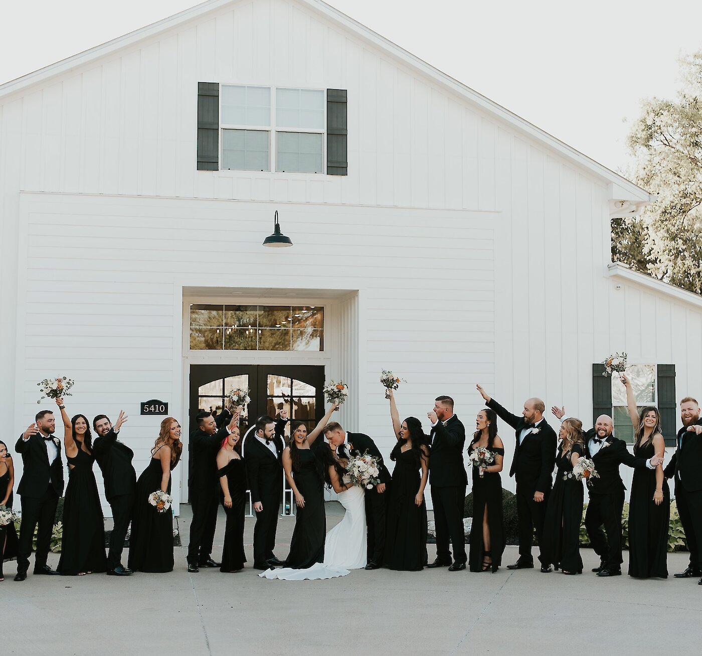 Large, celebratory wedding party portrait of the bride, groom, and ten attendants posing outside a modern white barn venue. The entire party wears matching black formal attire, cheering with raised arms.