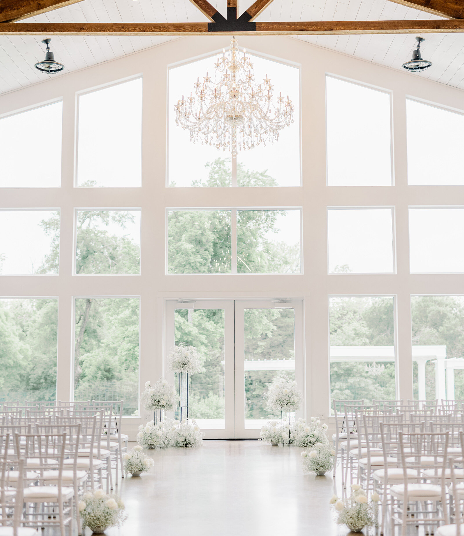 Sunny wedding portrait of the couple embracing on a white bridge overlooking a pond at a farmhouse venue. The groom wears a tan suit, and the bride wears an off-the-shoulder ballgown, with a white barn and silver silo visible in the background.