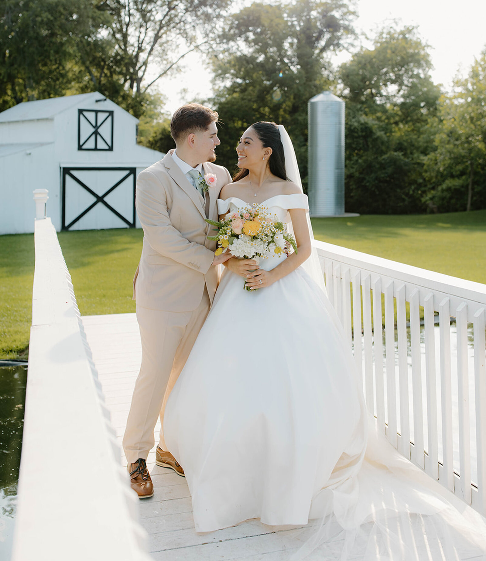 Bright indoor wedding ceremony in a spacious white venue with a high vaulted ceiling, exposed wooden beams, and a dramatic wall of windows behind the altar. Guests are seated in white chairs lining the aisle, facing the couple and wedding party.