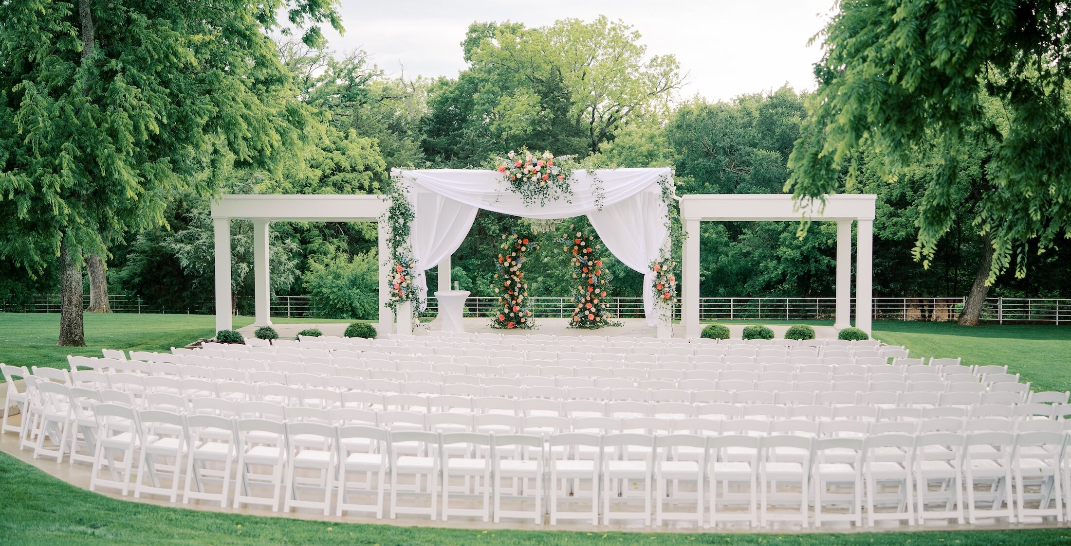 An outdoor wedding ceremony set up with rows of white chairs facing a white modern arbor draped with fabric and decorated with pink and coral flowers.