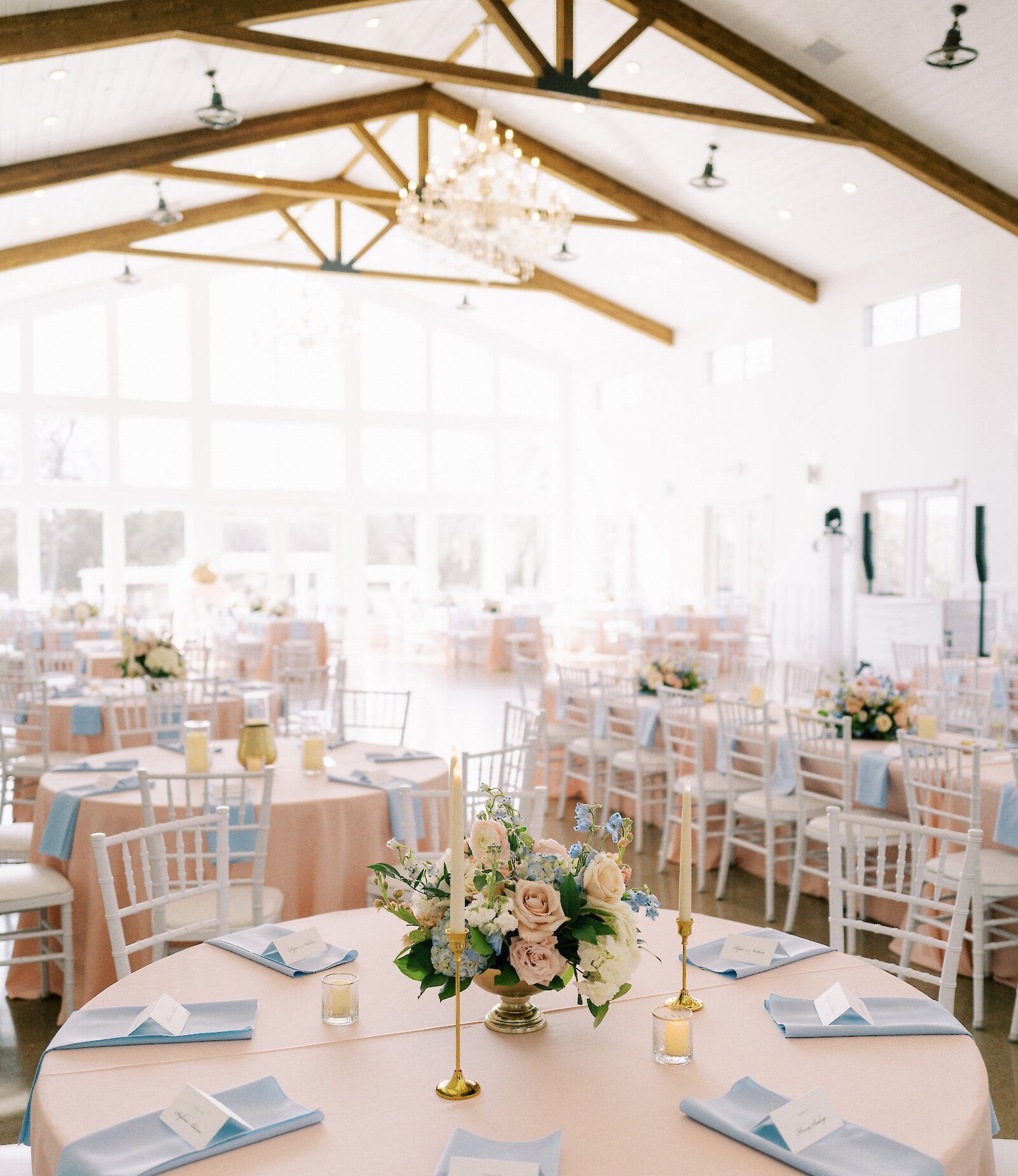 Bright wedding reception hall interior with a high white vaulted ceiling and exposed wooden trusses. Tables are set with blush pink tablecloths, light blue napkins, white chiavari chairs, and delicate centerpieces featuring pink and blue flowers.