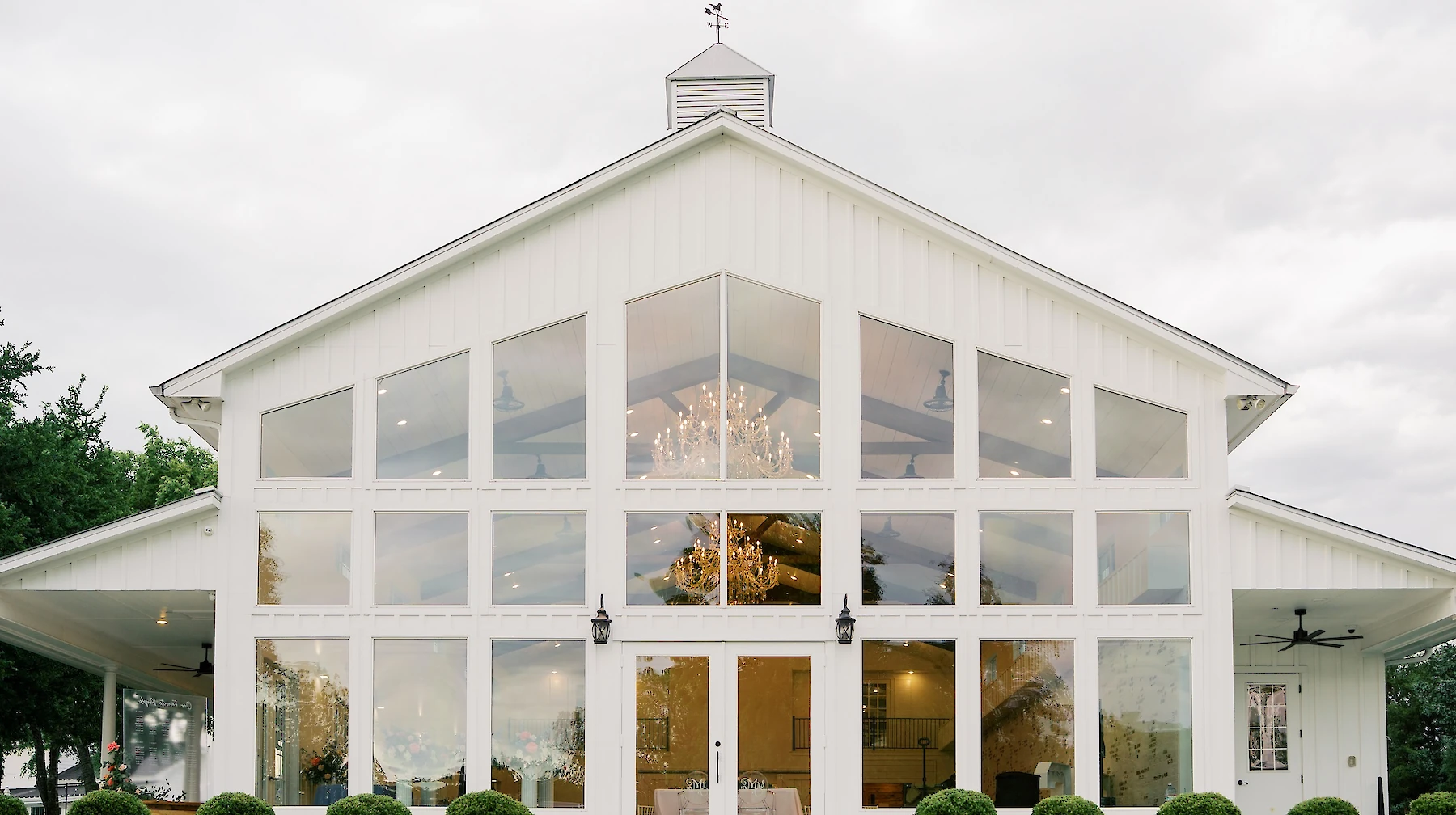 Grand exterior of a modern white barn wedding venue featuring a wall of tall, segmented glass windows under a high gable roof with a cupola. The interior chandeliers are visible, and the building is set behind steps leading up from a manicured green lawn.
