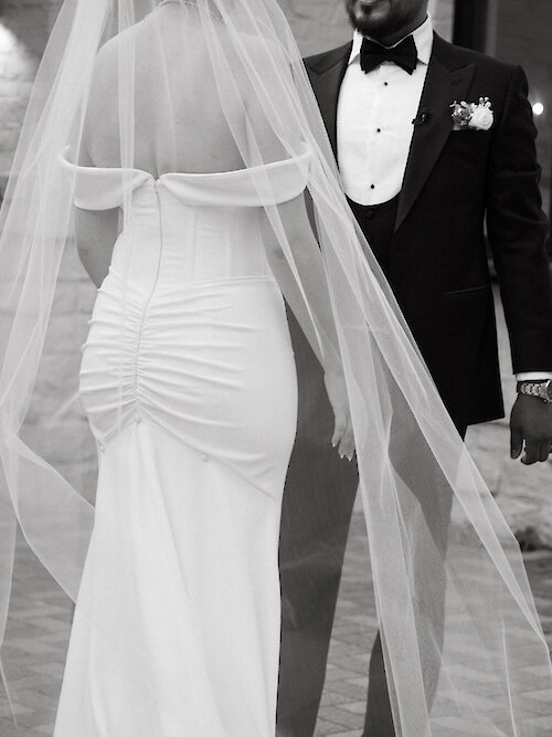 Black and white close-up wedding portrait highlighting the bride's figure-hugging off-the-shoulder gown with ruching and the groom's classic black tuxedo with a vest. The long veil drapes between the couple as they stand close together.