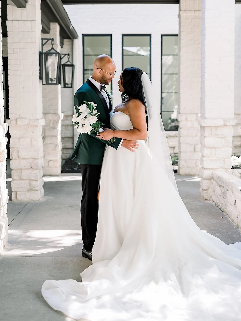 Intimate wedding portrait of a Black couple embracing and gazing at each other on a stone patio with white brick pillars. The groom wears a dark green tuxedo jacket, and the bride wears a strapless white satin ballgown with a flowing train.